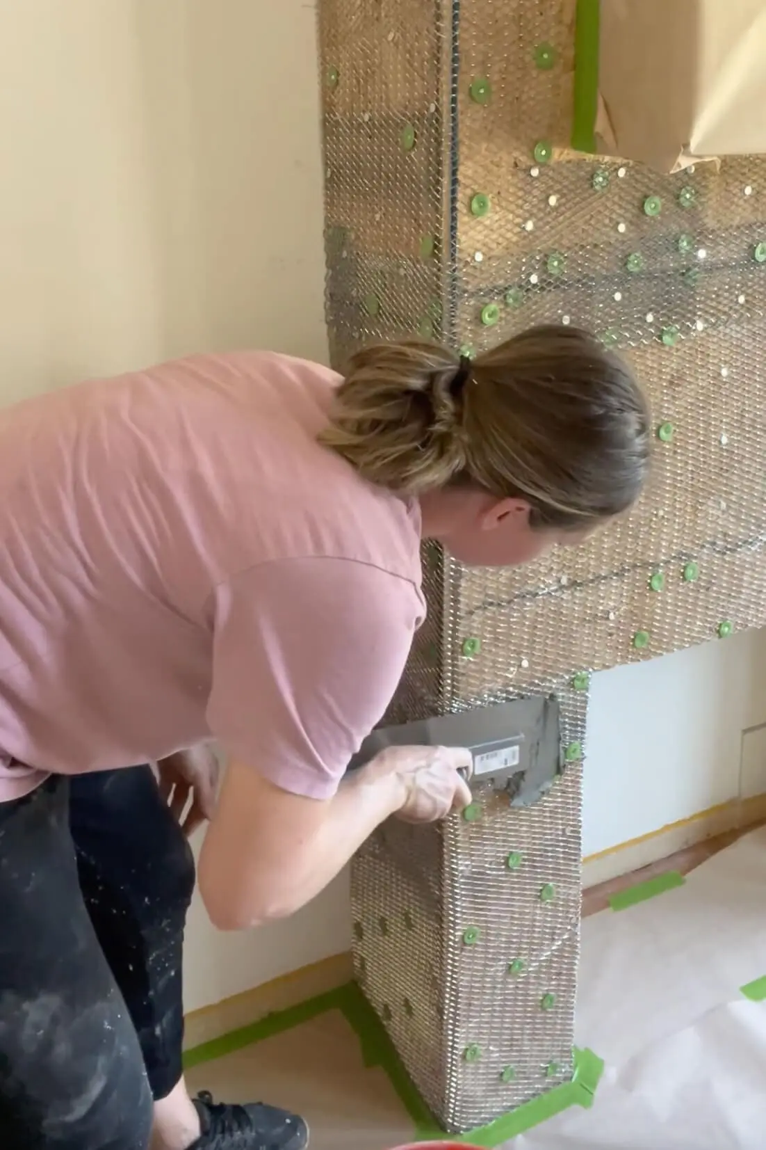 Woman applying mortar over metal lath on a framed fireplace structure during the early stages of a DIY stone fireplace build.